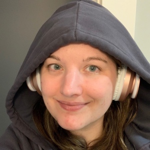 Photo of Robyn's face, a 35-year-old white woman, wearing a blue-gray hood and pink headphones, smiling and wearing no makeup.