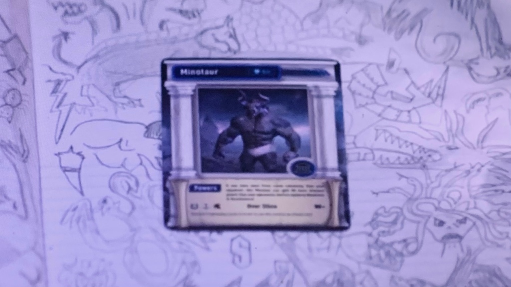 A screenshot from the show, zoomed in on young Percy's notebook. The page is full of pencil drawings of mythological creatures. A Mythomagic playing card lies on top, featuring the Minotaur - a man with a bull's head - wearing white underwear.